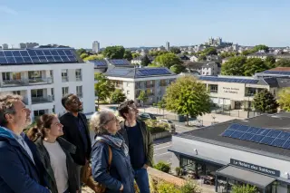 Urban buildings in Le Mans with solar panels on roofs and a group of people looking at them under clear sunny sky.